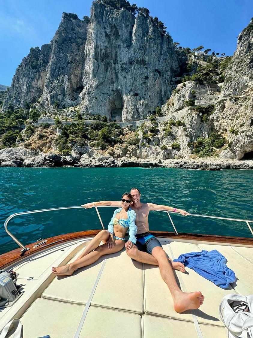 Linda and Collin on the bow of a boat, cliffs and turquoise water behind them