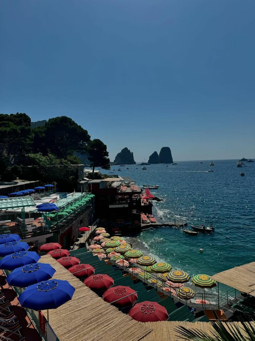 Capri beach-club umbrellas in rows, the Faraglioni rocks out in the water
