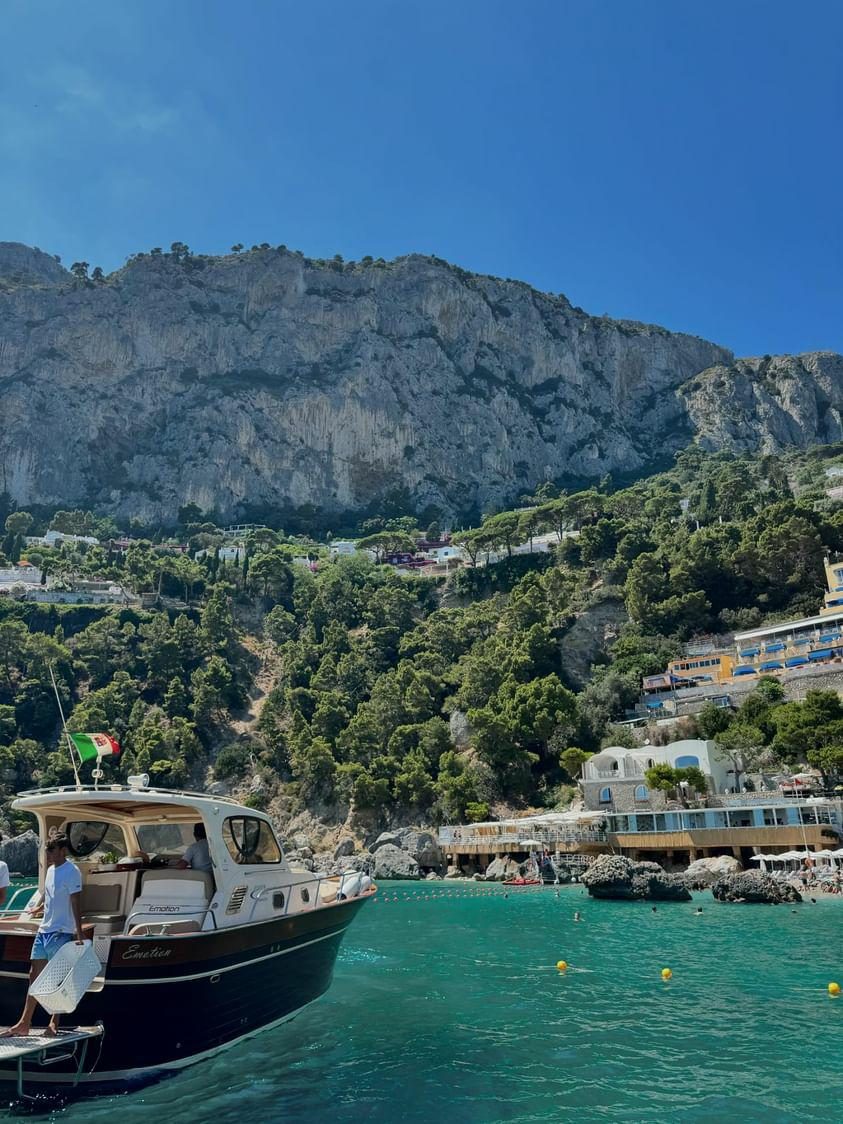 A boat off a rocky beach club, Italian flag on the stern, cliffs behind
