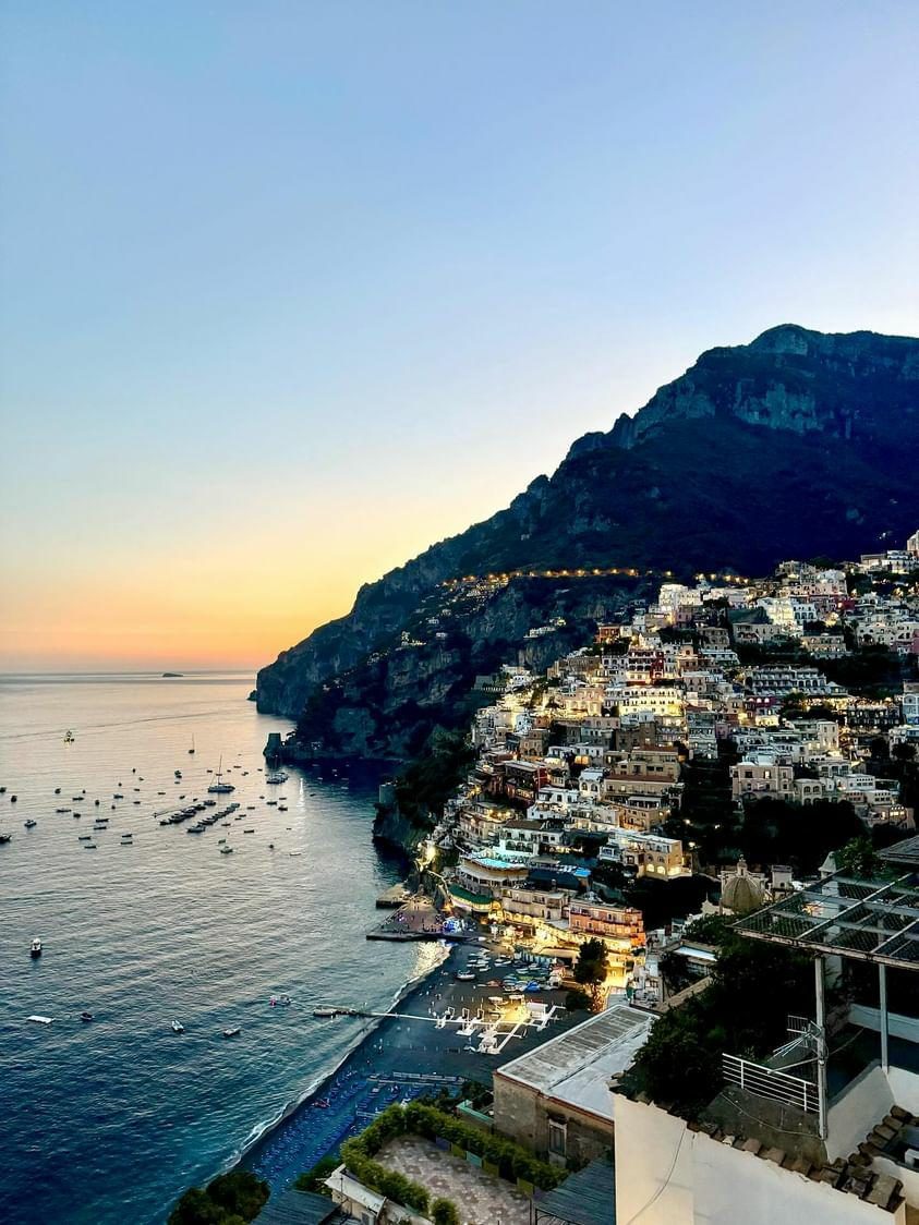 Positano at dusk, the hillside lit, boats in a darkening bay