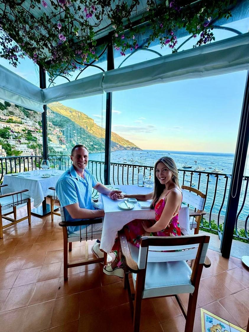 Linda and Collin holding hands at a terrace table, Positano and the bay behind them
